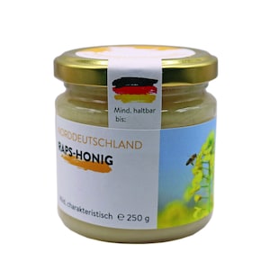 May include: A glass jar of light yellow honey with a white label that says "Norddeutschland Raps-Honig" and a German flag. The label also says "Mind. haltbar bis:" and "e 250 g".