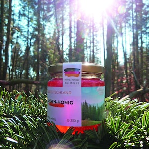 May include: A glass jar of honey with a red label that says "Tannen-Honig" and a picture of a forest. The label also says "Made in Germany" and "Min. halbar bis: 31.09.23". The jar is sitting on a bed of green pine needles.