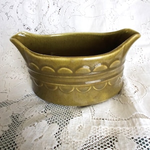 May include: A vintage, olive-green ceramic gravy boat. The boat has a scalloped design around the base and a curved rim with a spout on each end. The gravy boat is sitting on a white lace tablecloth.