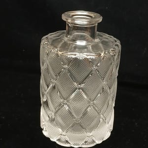 May include: A clear glass bottle with a diamond pattern on the body. The bottle has a cylindrical shape with a narrow neck and a wide, flat base. The glass is transparent, allowing light to pass through.