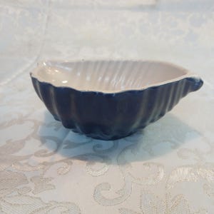 May include: A small, ceramic dish with a seashell design. The dish has a navy blue exterior and a white interior with a ribbed texture. The dish is oval-shaped and has a small handle on one side.