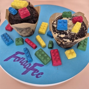 Könnte beinhalten: Zwei Schokoladenmuffins in Papierförmchen, verziert mit bunten, essbaren Baustein-Süßigkeiten. Die Muffins stehen auf einem blauen Teller mit dem Text "FonaaFee" in Schreibschrift. Die Süßigkeiten sind rot, gelb, grün und blau.