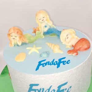 Könnte beinhalten: Ein weißer Kuchen, verziert mit Fondant-Meerjungfrauenfiguren, Muscheln und einem Seestern. Der Kuchen ist in Blau glasiert und trägt die Aufschrift "FondaFee" in blauer Glasur.