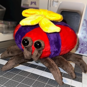 Puede incluir: Una araña de peluche con cuerpo rojo, rayas moradas y patas marrones. La araña tiene ojos grandes y negros y un lazo amarillo en la cabeza. El juguete está sobre una esterilla de corte gris.