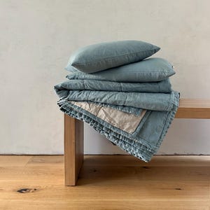 May include: A stack of blue linen pillows and a blue and white quilted throw blanket on a wooden bench.