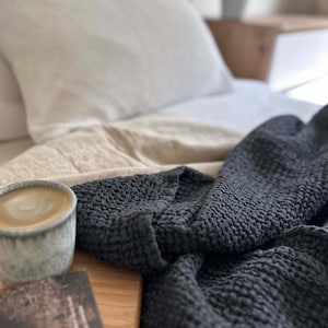 Peut inclure: Gros plan sur une couverture en tissu gaufré gris foncé sur un lit avec un oreiller blanc et une tasse de café.