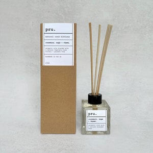 May include: A clear glass bottle with a black lid and wooden sticks, filled with a light yellow liquid. The bottle has a label that reads "pru. rosemary, sage thyme. aromatic oils with a natural blended base". The bottle is sitting on a white surface next to a brown cardboard box with a similar label.