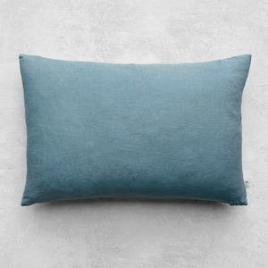 Puede incluir: Una almohada rectangular azul claro con una superficie texturizada. La almohada es de color liso y parece estar hecha de lino o un tejido similar. La almohada está sobre una superficie gris claro.