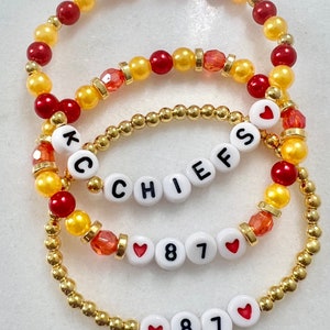 Könnte beinhalten: Drei Perlenarmbänder mit roten und goldenen Perlen. Das obere Armband hat das Wort "CHIEFS" in weißen Perlen geschrieben. Das mittlere Armband hat ein Herz, die Zahlen 8 und 7 und ein weiteres Herz. Das untere Armband hat ein Herz, die Zahlen 8 und 7 und ein weiteres Herz.