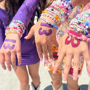 Op de afbeelding: Drie vriendinnen dragen kleurrijke kralenarmbanden met woorden en cijfers. De armbanden zijn gestapeld op hun polsen. De vriendinnen hebben het nummer 33 met een paarse stift op hun handen geschreven.