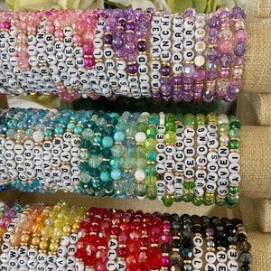 Op de afbeelding: Een stapel kleurrijke kralenarmbanden met verschillende woorden die op elk armband zijn gespeld. De woorden omvatten "Say Don't Tell", "Clean", "Style", "Love", "Sparks", "Ours", "Enchanted", "Reputa", "Labyrinth", "Get", "OMG", "Tim", "CGAW", "Uris", "Bo", "But", "Song", "Seven", "All Too", "Fearless", "Flower", "White", "Esse", "Love", "Street", "Long", "Last", "Jours", "Live", "Dear", "Almost", "Sn", "19", "Eve", "Per", "Sweet", "Free", "Daylight", "Swift", "Per", "My", "Su", "U", "R".