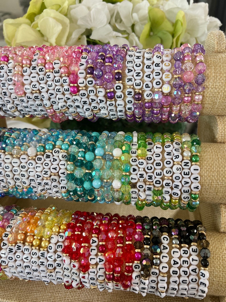 Op de afbeelding: Een stapel kleurrijke kralenarmbanden met verschillende woorden die op elk armband zijn gespeld. De woorden omvatten "Say Don't Tell", "Clean", "Style", "Love", "Sparks", "Ours", "Enchanted", "Reputa", "Labyrinth", "Get", "OMG", "Tim", "CGAW", "Uris", "Bo", "But", "Song", "Seven", "All Too", "Fearless", "Flower", "White", "Esse", "Love", "Street", "Long", "Last", "Jours", "Live", "Dear", "Almost", "Sn", "19", "Eve", "Per", "Sweet", "Free", "Daylight", "Swift", "Per", "My", "Su", "U", "R".