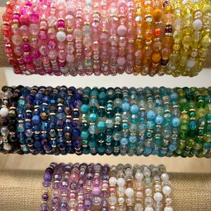 Op de afbeelding: Een stapel kleurrijke kralenarmbanden in tinten roze, oranje, blauw, groen en paars. De armbanden zijn gemaakt van kleine, ronde kralen en zijn gerangschikt in een verloop.