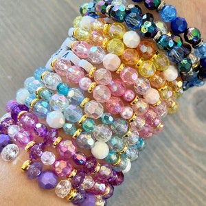 Op de afbeelding: Een stapel kleurrijke kralenarmbanden met iriserende glazen kralen en gouden accenten. De armbanden zijn gerangschikt in een regenboogpatroon, beginnend met paars en eindigend met blauw.