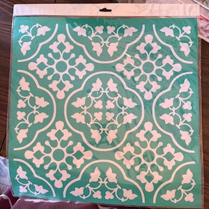 May include: A square stencil sheet featuring a repeating white floral and geometric pattern on a teal background. The design consists of interconnected quatrefoil shapes and stylized leaf motifs, all enclosed within a clear plastic protective sleeve.