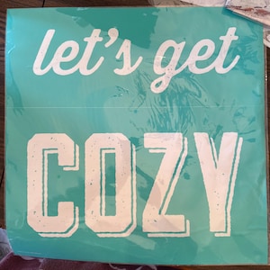 May include: A square teal sign featuring the white text let's get in a cursive script font on the top half and the word COZY in a large white block letter font on the bottom half. The letters have a distressed texture throughout the design.
