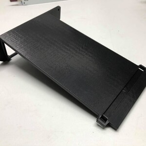 May include: A black 3D printed phone stand with a slot for charging cables. The stand is angled to hold a phone upright.
