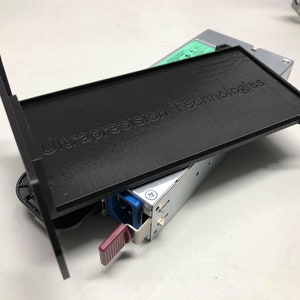 May include: A black plastic cover with the text "Ultraprecision Technologies" printed on it. The cover is partially open, revealing a silver power supply unit with a blue connector.