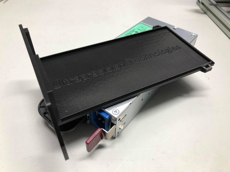 May include: A black plastic cover with the text "Ultraprecision Technologies" printed on it. The cover is partially open, revealing a silver power supply unit with a blue connector.