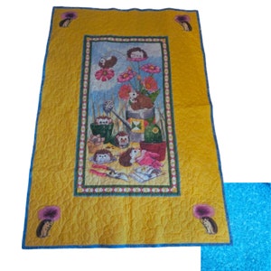 Puede incluir: Una manta acolchada amarilla con un borde azul y un panel central con una ilustración colorida de erizos en un entorno de jardín. El panel incluye una regadera, flores y herramientas de jardinería.