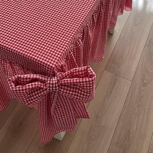 May include: A red and white gingham tablecloth with a ruffled skirt and large bow details. The table covering is draped over a table with white legs, creating a classic, country-style aesthetic.