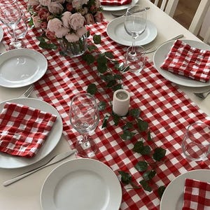 Puede incluir: Una mesa puesta con un camino de mesa a cuadros rojos y blancos, platos blancos y servilletas a juego. Copas de vino, un centro de mesa floral y una vela completan la disposición. La mesa está lista para una comida.