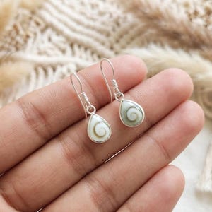 Op de afbeelding: Een paar zilveren oorbellen met druppelvormige Shiva-oog schelpinleg. Elke oorbel heeft een gepolijste zilveren zetting met een spiraalpatroon op de schelp, hangend aan een eenvoudige zilveren haak voor gaatjes in de oren.