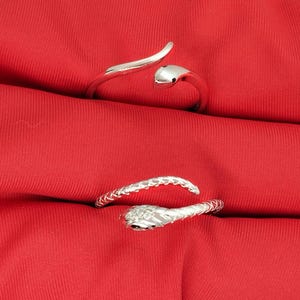 Könnte beinhalten: Zwei silberfarbene Armbänder in Schlangenform liegen auf einem leuchtend roten Stoff. Die Armbänder weisen detaillierte Schlangenkopfdesigns und strukturierte Muster auf. Der Schmuck wird vor einem einfarbigen Hintergrund präsentiert.