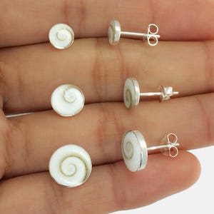 May include: Three pairs of stud earrings. Each earring features a white, circular design with a spiral pattern, set in a silver-colored metal. The earrings are displayed on a light background.