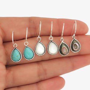 Peut inclure: Une collection de six boucles d'oreilles en forme de goutte d'eau avec des cerceaux argentés. Les boucles d'oreilles présentent diverses incrustations de pierres, dont de la turquoise, de la nacre blanche et de l'ormeau. Chaque boucle d'oreille a une bordure argentée en forme de corde.