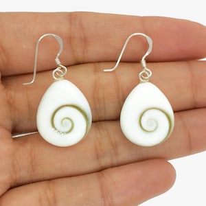 May include: A pair of white teardrop earrings with a silver hook. Each earring has a green spiral design. The earrings are displayed on a hand.