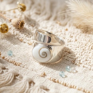 May include: A silver ring featuring a white, spiral-patterned central stone. The ring sits on a textured, cream-coloured surface, with dried flowers and small blue beads. The ring's design is contemporary and stylish.