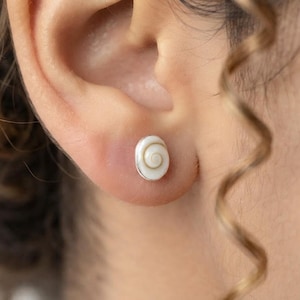 Op de afbeelding: Een kleine ovale oorsteker met een natuurlijke witte schelp met een bruin spiraalpatroon in het midden van het oppervlak. De schelp is gezet in een eenvoudige zilverkleurige metalen rand, wat zorgt voor een compact en rond ontwerp.