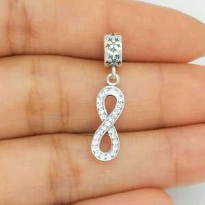 May include: A silver charm with an infinity symbol pendant, encrusted with clear crystals. The top section features snowflake designs. This jewellery piece is designed to be attached to a bracelet or necklace.