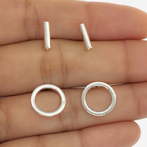 May include: A set of silver stud earrings. The set includes two small, straight bar earrings and two circular hoop earrings. The earrings are displayed on a light background.