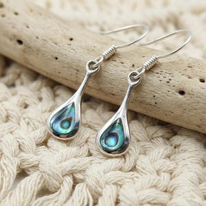 Peut inclure: Une paire de boucles d'oreilles pendantes argentées dotées d'une monture en forme de goutte avec une incrustation de coquille d'ormeau irisée. Les boucles d'oreilles ont une attache à crochet simple et une finition métallique polie aux reflets bleus et verts.
