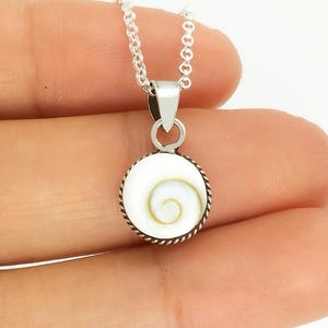 Puede incluir: Un collar de plata con un colgante circular. El colgante presenta un fondo blanco con un diseño en espiral en el centro. El colgante está enmarcado por un borde trenzado en forma de cuerda. El collar está sujeto por una mano.