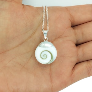 May include: A silver necklace with a pendant featuring a white, round shell design and a green spiral pattern. The pendant is suspended from a silver chain. The necklace is shown on a person's open palm.