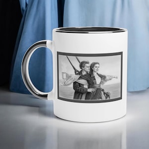 May include: White ceramic mug with a black rim and silver handle. The mug features a black and white pencil drawing of a couple on a ship, with the woman's arms outstretched. The image is framed in black.