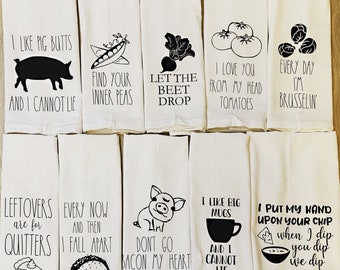 May include: A collection of white kitchen towels with black graphic designs and text. Designs include a pig, peas, a beet, tomatoes, Brussels sprouts, a pie, a taco, a pig, a mug, and a bowl with chips. Text includes humorous phrases.