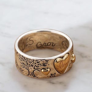 Può includere: Un anello bicolore con interno dorato ed esterno argentato. L'anello presenta un disegno ad albero, cuori e la parola "love" su un accento a forma di cuore. Le parole "JES grow" sono incise sull'anello.