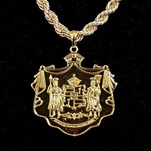 Könnte beinhalten: Goldfarbene Halskette mit Anhänger und detailliertem Wappen-Design. Der Anhänger hängt an einer dicken, gedrehten Seilkette. Das Wappen zeigt Figuren, eine Krone und Text. Der Hintergrund ist schwarz.
