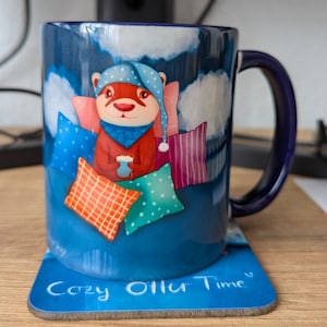 May include: A blue ceramic mug featuring a cartoon raccoon wearing a blue and white sleep cap and holding a cup. The mug has a white handle and the text "Cozy Ollie Time" on the bottom.