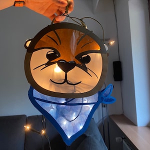 May include: A paper lantern shaped like an otter's face with a blue bandana. The lantern is lit with white fairy lights.