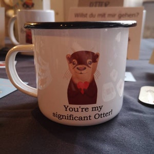 Könnte beinhalten: Eine weiße Emailletasse mit schwarzem Rand und Henkel. Die Tasse zeigt eine Comic-Otter, die eine rote Fliege trägt und den Text "You're my significant Otter!"