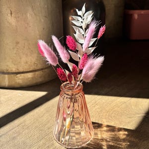 May include: A small, pink glass vase with a fluted design holds a bouquet of dried flowers. The arrangement includes pink and red fluffy stems, and white, leafy sprigs. The vase sits on a wooden surface, with sunlight casting shadows.