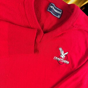 Peut inclure: Un pull rouge vif à col en V avec des poignets et un col côtelés. Le pull présente un motif d'aigle brodé avec le mot "CRAFTSMAN" en dessous. Une étiquette indique "SETH ROBERTS".