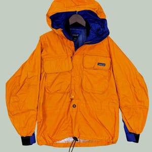 Puede incluir: Una chaqueta con capucha de color naranja brillante con un forro interior y capucha en azul oscuro. La chaqueta incluye dos grandes bolsillos delanteros en el pecho, un cierre central de botones y un dobladillo con cordón. Etiqueta Patagonia.
