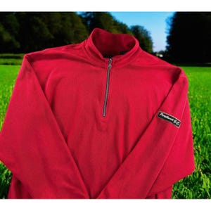 May include: A vibrant red long-sleeve pullover with a quarter-zip closure and a stand-up collar. The sleeve features a small black patch with white text. The pullover is made of a soft material, perfect for cooler weather.