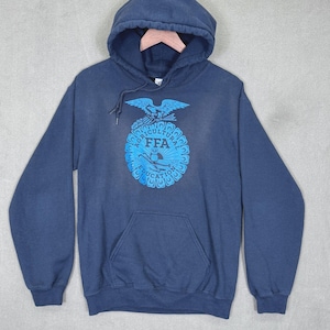 May include: A navy blue hooded sweatshirt featuring a front kangaroo pocket and long sleeves. The center of the chest displays a bright blue graphic emblem with an eagle, an owl, and the text Agricultural Education FFA in a circular design.